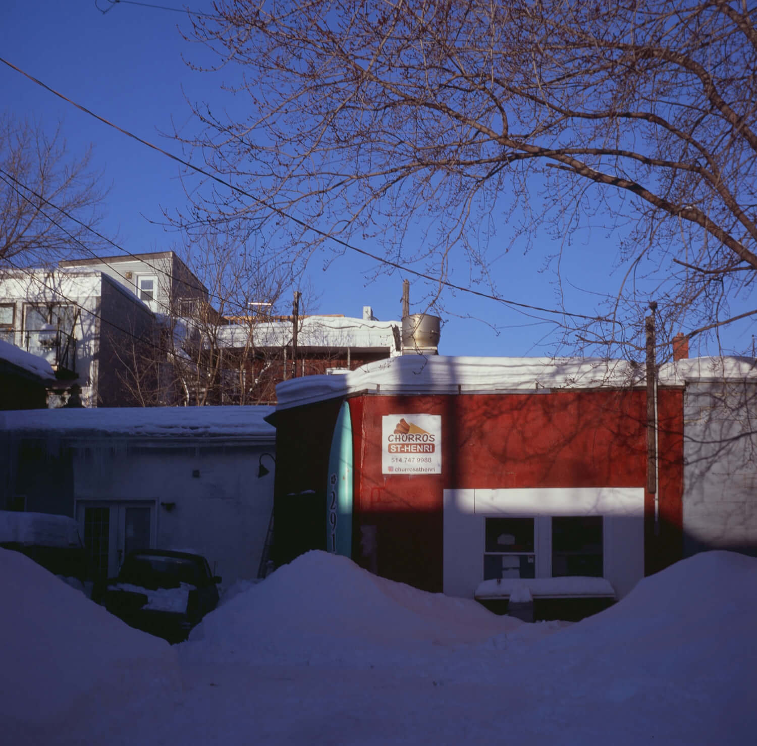 An image of a snow-covered driveway of a churro shop. The sun is hitting a red building at an angle, leaving the snow in shadow. 