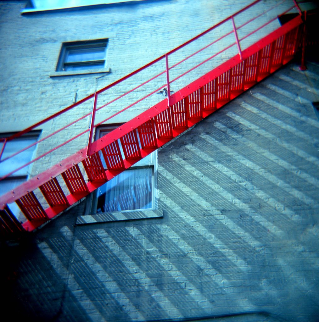 a red fire escape casting shadows on the side of the building it serves
