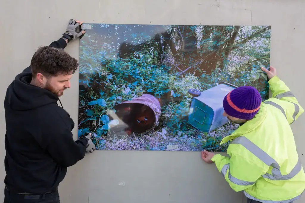 Two men line up a 1.5 metre print on a building site hoarding. The print shows a Henry vacuum cleaner peering out from inside a rusted metal drum dumped in woods.