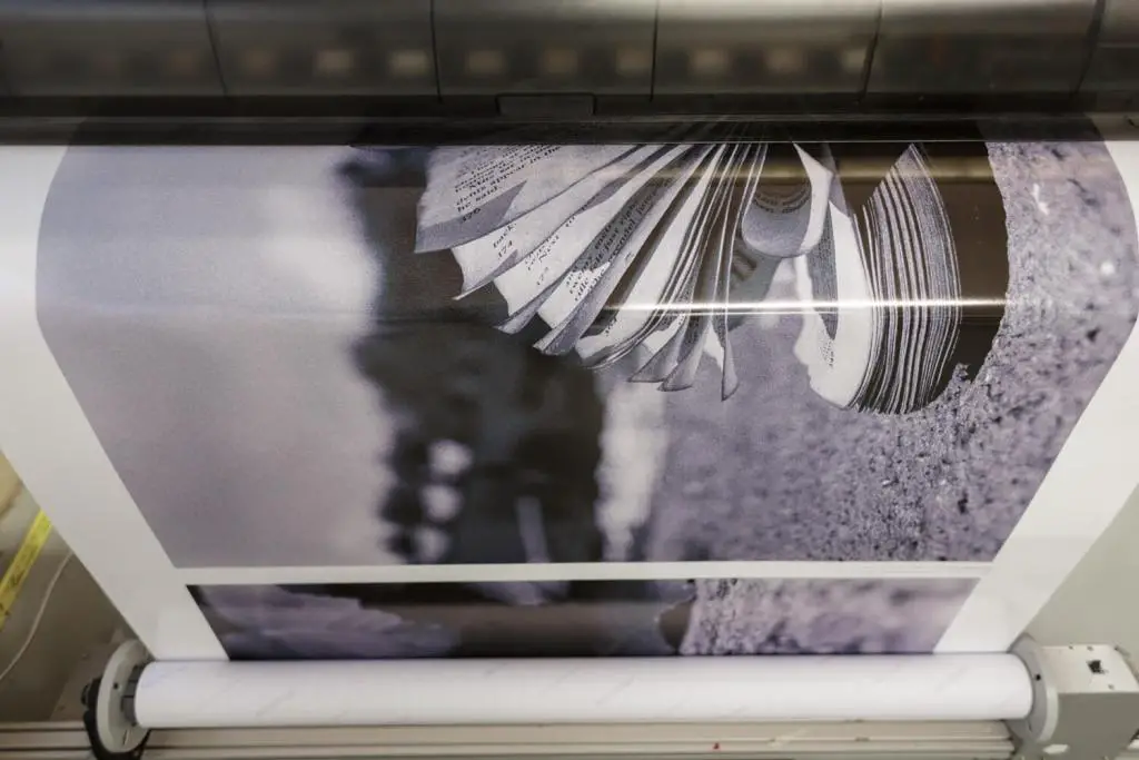 A black and white large-format print showing the pages of a discarded book is seen coming out of an inkjet printer.