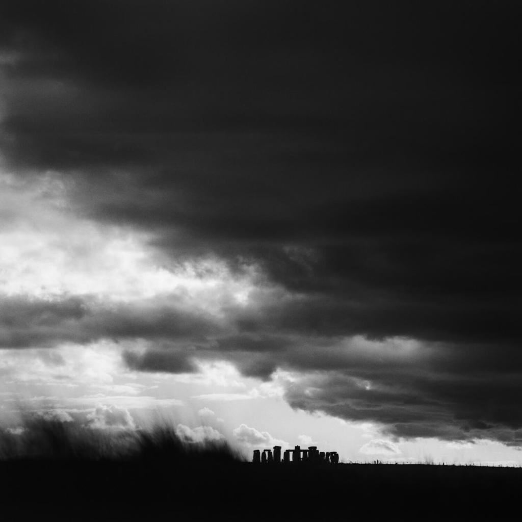 Black and white photo of Stonehenge silhouetted and seen from a distance with very dark storm clouds building up in the sky.