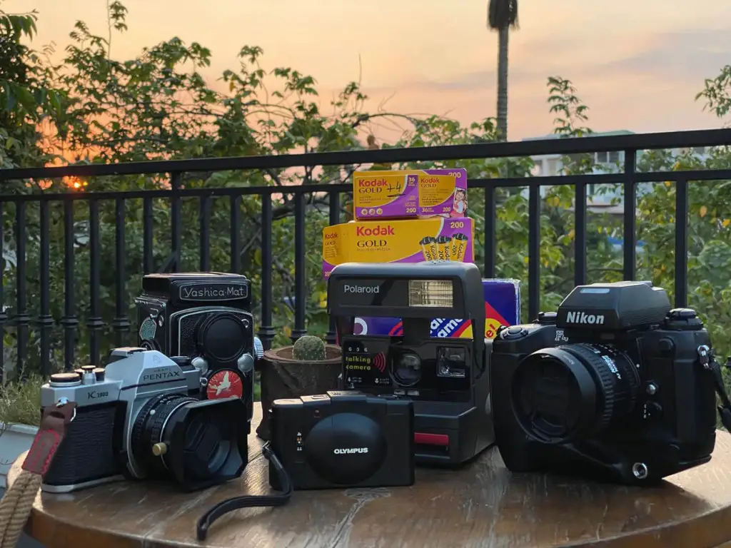 The five cameras I used to take pictures with, on a table in a nice outdoor coffee shop, the cameras are next to each other and they are - from left to right - Pentax K1000, Yashica Mat, Olympus XA2, Polaroid 636, and Nikon F4S