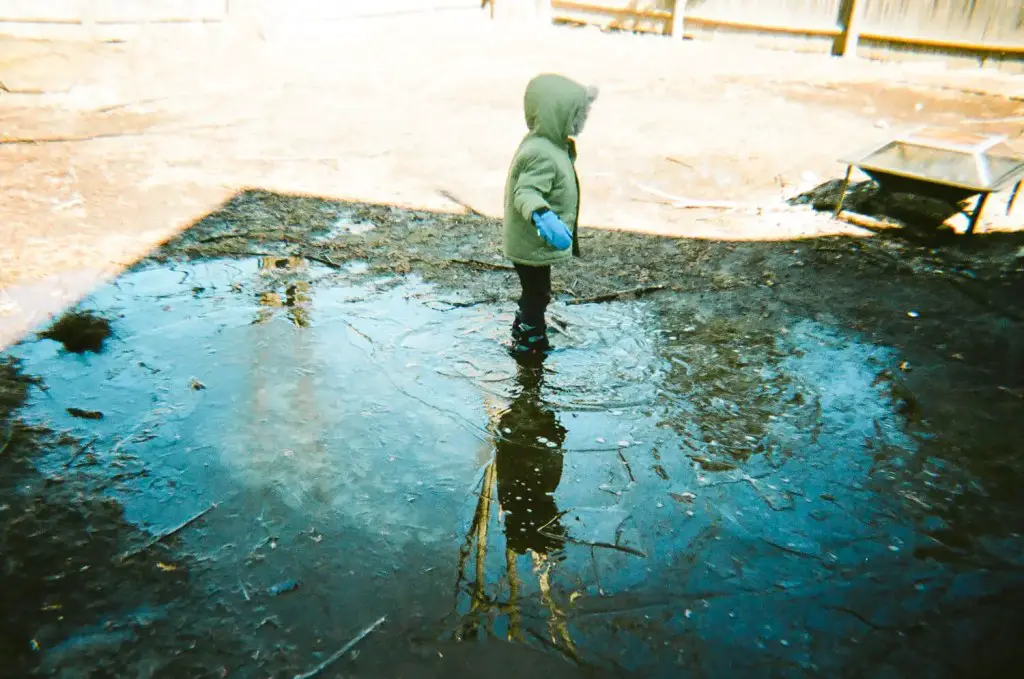 Profile of child in green coat with blue mittens standing in a snowy puddle.  The blue sky is reflected in the puddle.  5 Frames of Kodak Portra 400 on Kodak M35 plastic camera - Kerry Constantino.  By